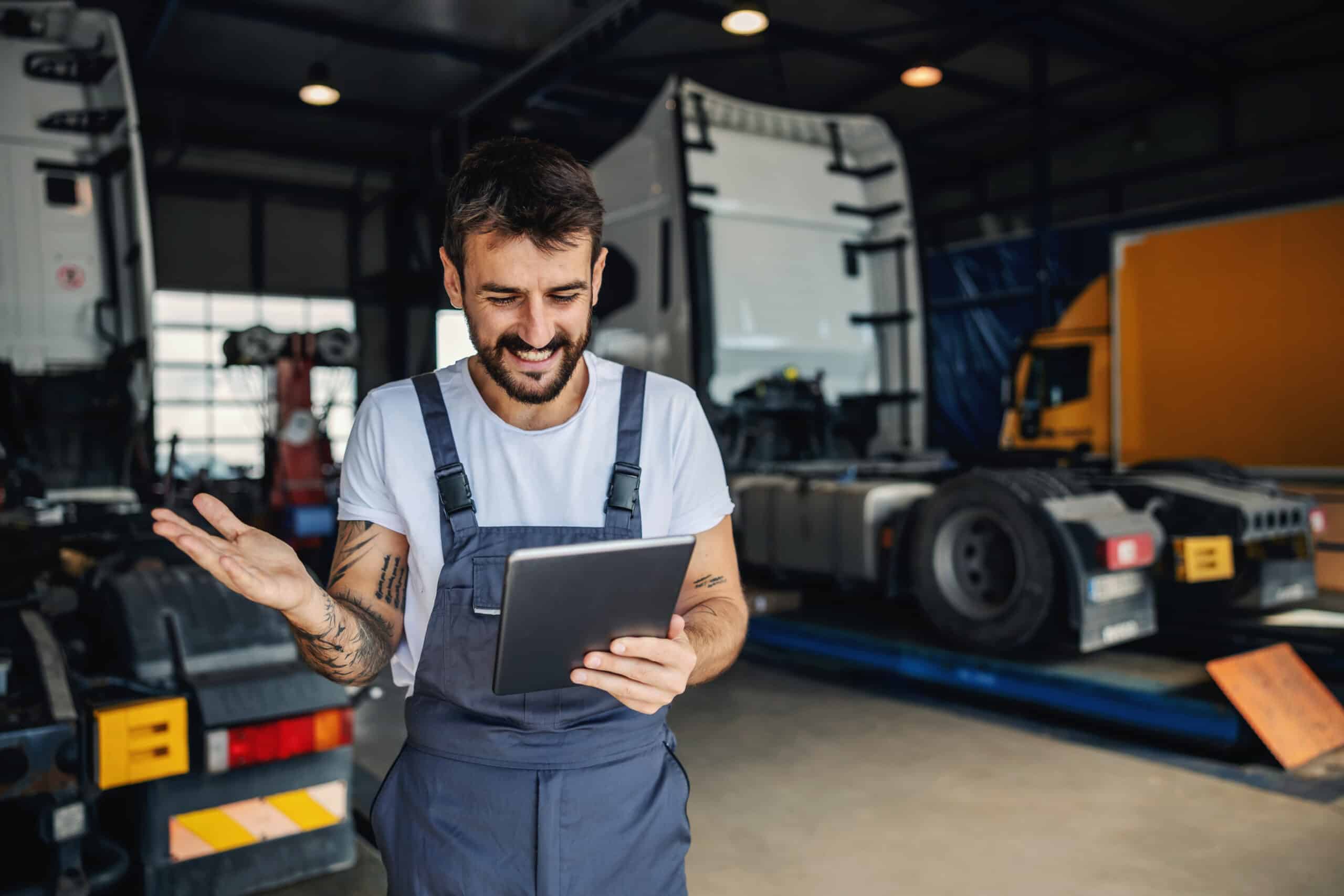 A smiling male mechanic in blue overalls and a white t-shirt, with visible arm tattoos, stands in a busy truck workshop. He is looking down at a digital tablet he holds, with one hand slightly raised as if explaining or reacting to information on the screen. In the background, large commercial trucks are visible, including one on a lift. This image illustrates a mechanic efficiently using a digital solution, like Autocheck, for vehicle inspections, job sheets, and managing fleet maintenance in a modern UK workshop setting.